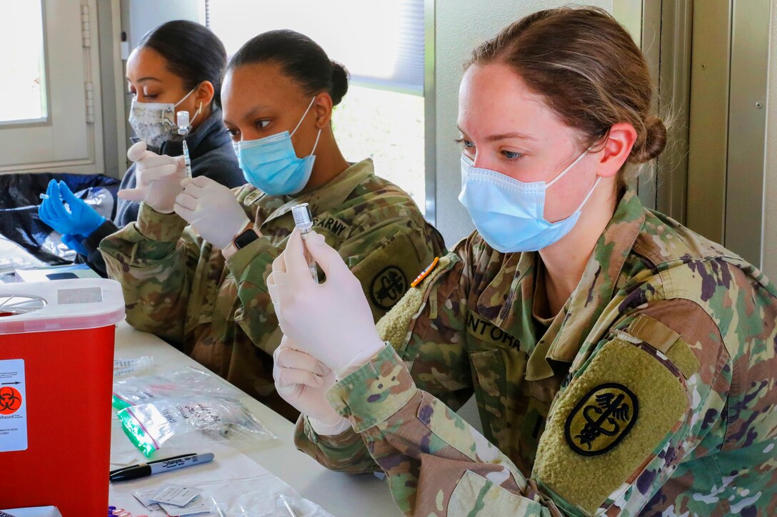 Two soldiers and a FEMA volunteer sit side by side at a table wearing face masks and gloves preparing vaccines.