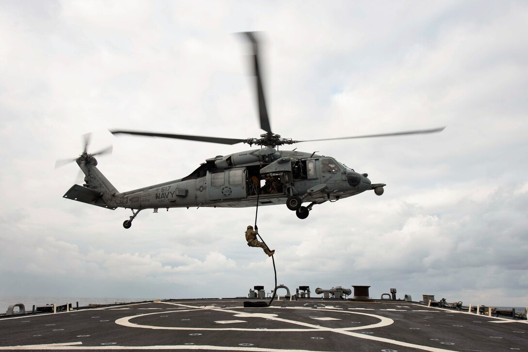 A sailor climbs down a rope from a helicopter hovering over a ship.