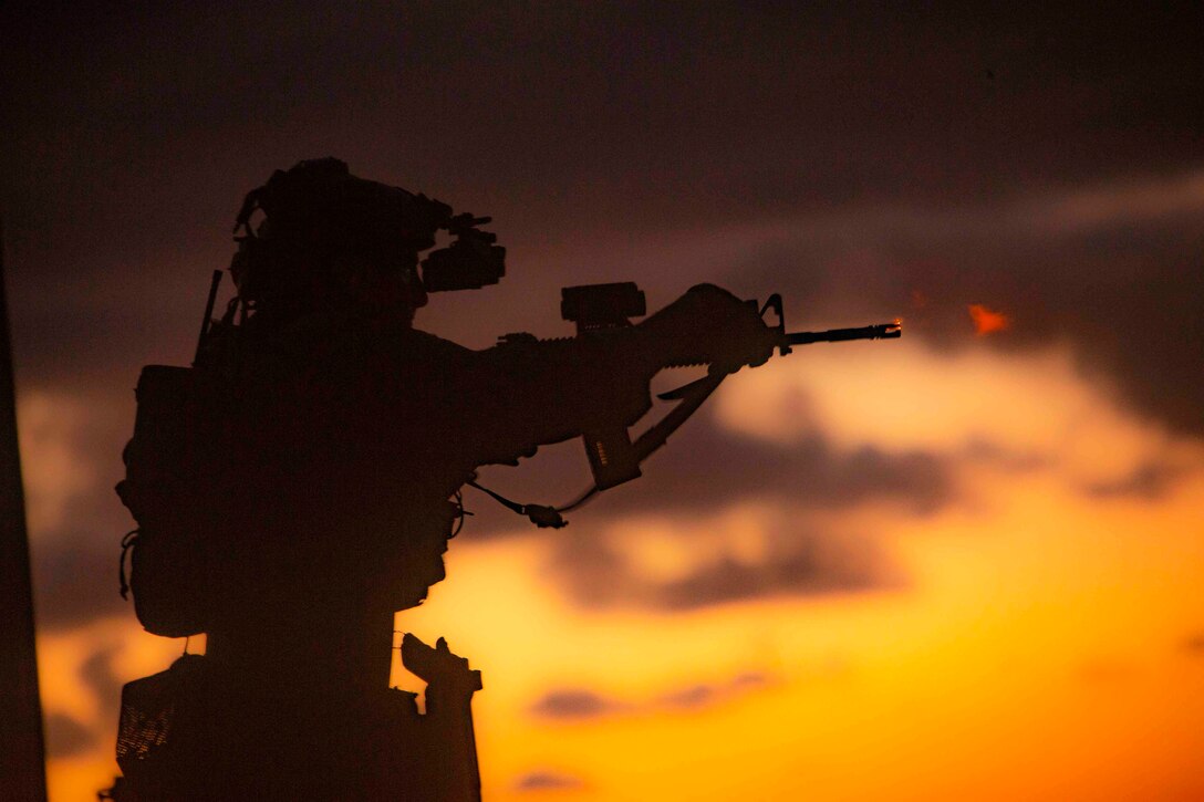 A service member shown in silhouette points a weapon toward a target.
