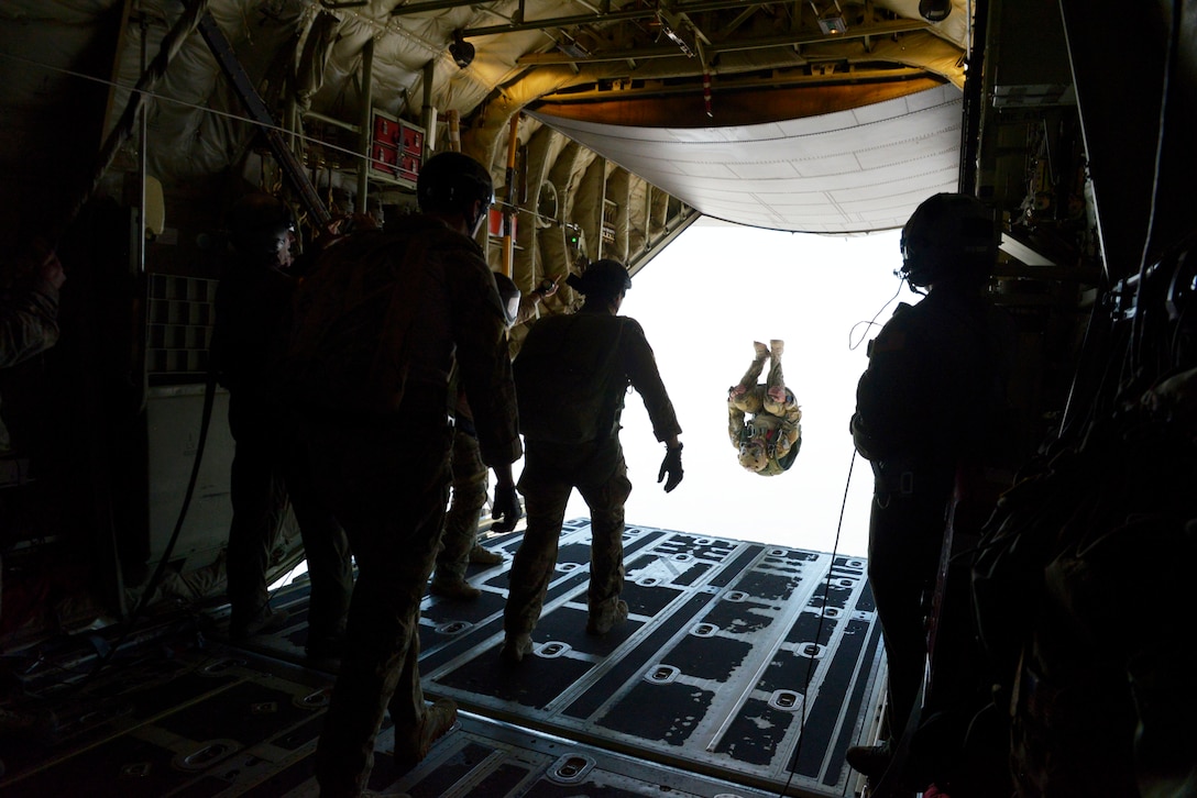 Several service members gather in the rear of an aircraft. Another has jumped out and appears to be suspended in the air.