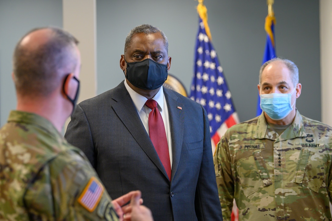 A man in a dark suit speaks with two other men in military uniforms.