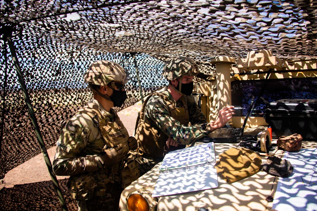 Two Marines examine a weapon system under camo netting.