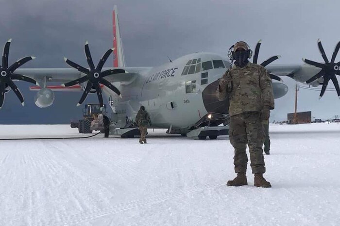 An airman stands on the ice with a large military aircraft behind him.