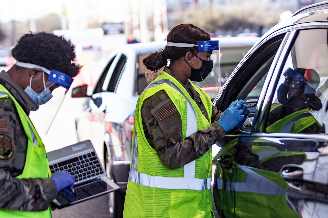 A female service member wearing a face mask and gloves stands at the driver's side window of a vehicle holding a syringe; another woman service member, who is also wearing a face mask and gloves, stands holding a laptop.