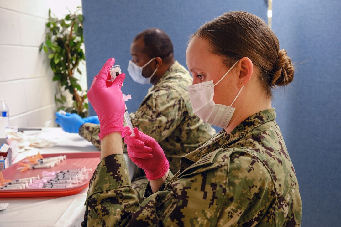 A female service member holds a syringe, which is inserted into a small bottle; a male service member seated nearby looks at something in his hands. Both are wearing face masks and gloves.
