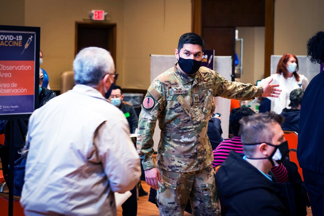 A service member wearing a face mask motions to an elderly man.