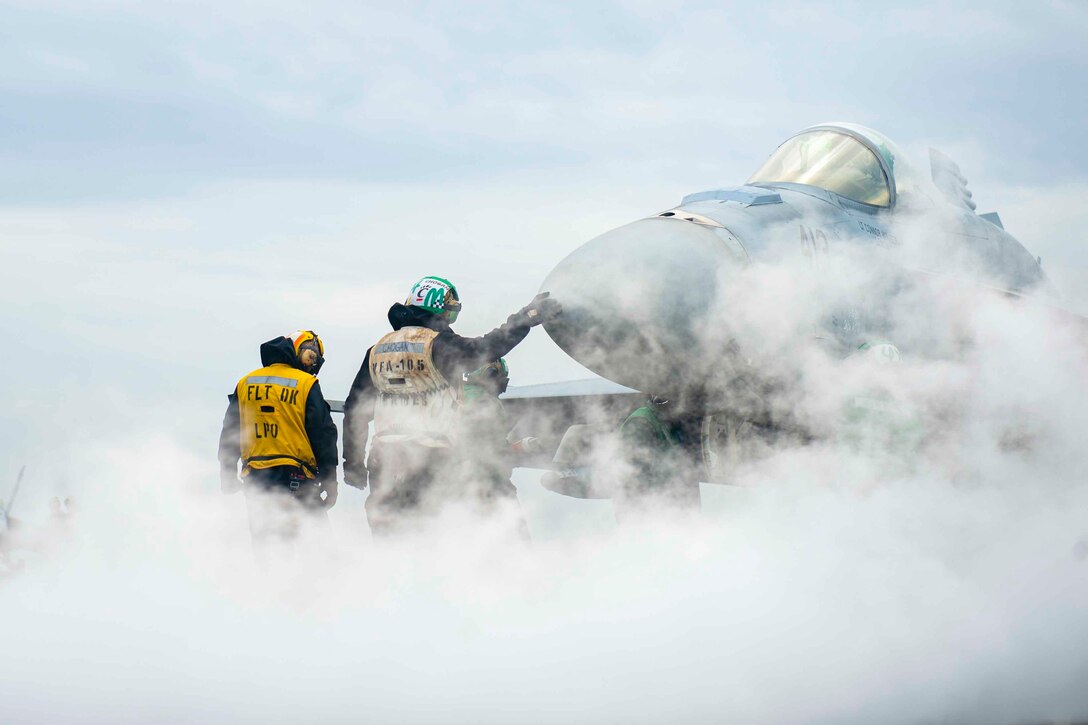 Sailors stand next to an aircraft as smoke surrounds them.