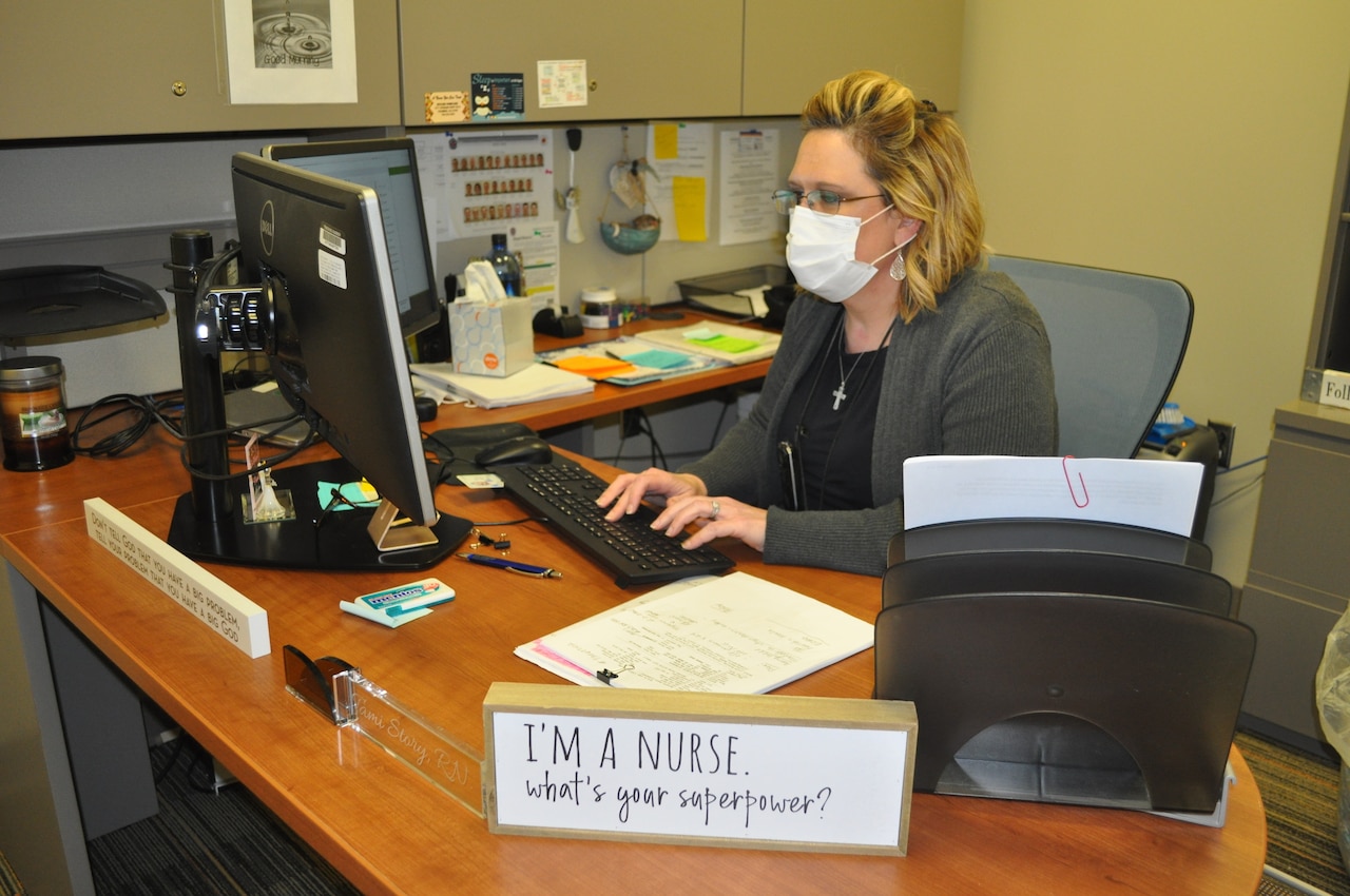 A female nurse in personal protective equipment works at her desk.