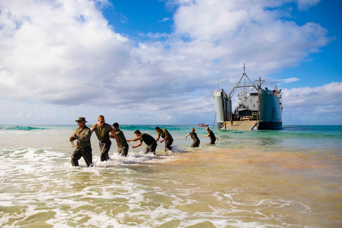 Marines stand on a beach in water up to their knees while pulling a rope connected to a large vessel.