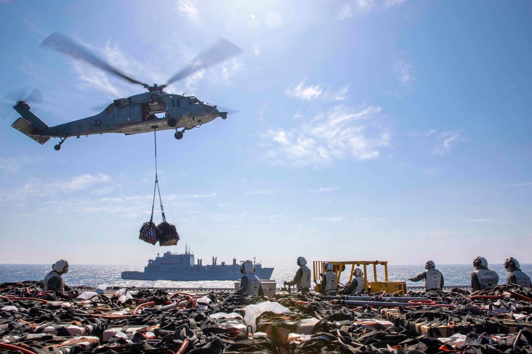 Marines stand on the deck of a ship as helicopter flies above.