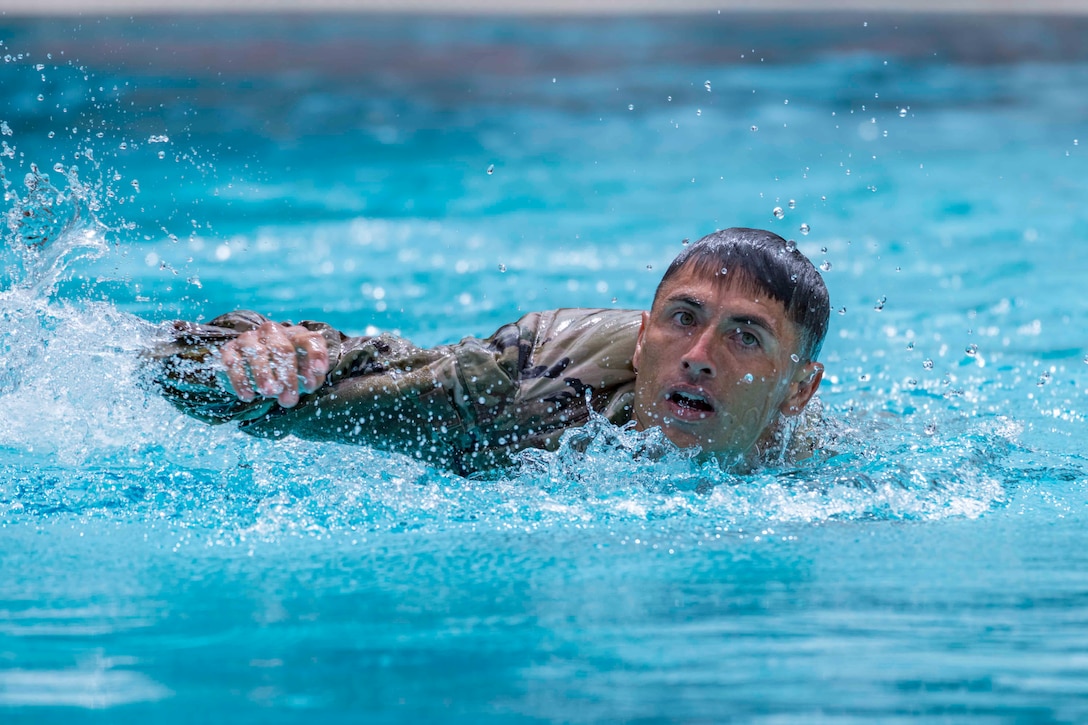 A soldier swims in a pool while wearing a military uniform.