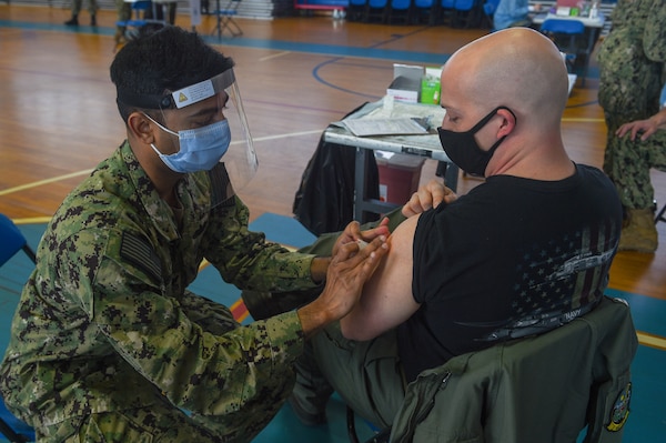 Hospital Corpsman 1st Class Sujit Rajendran, dental leading petty officer for Pre-Commissioning Unit (PCU) John F. Kennedy (CVN 79), administers a Pfizer COVID-19 vaccine to Lt. Matthew Hinkson, pilot for Helicopter Sea Combat (HSC) Squadron 11.
