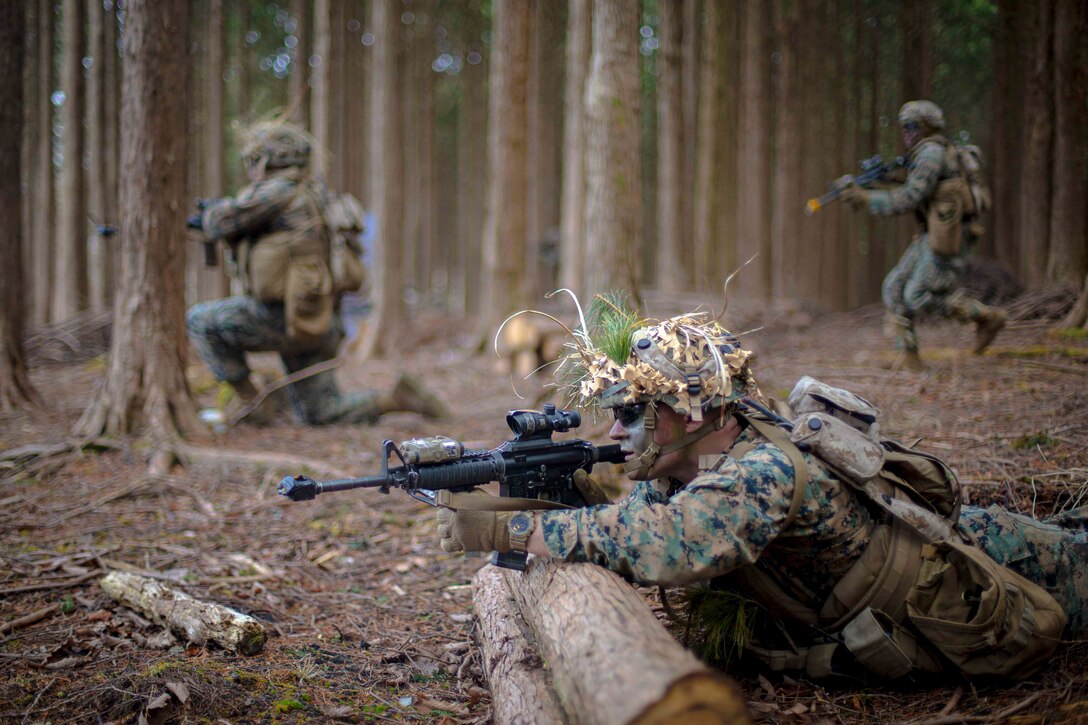 A Marine lies on the ground pointing a weapon as two others move through trees behind.