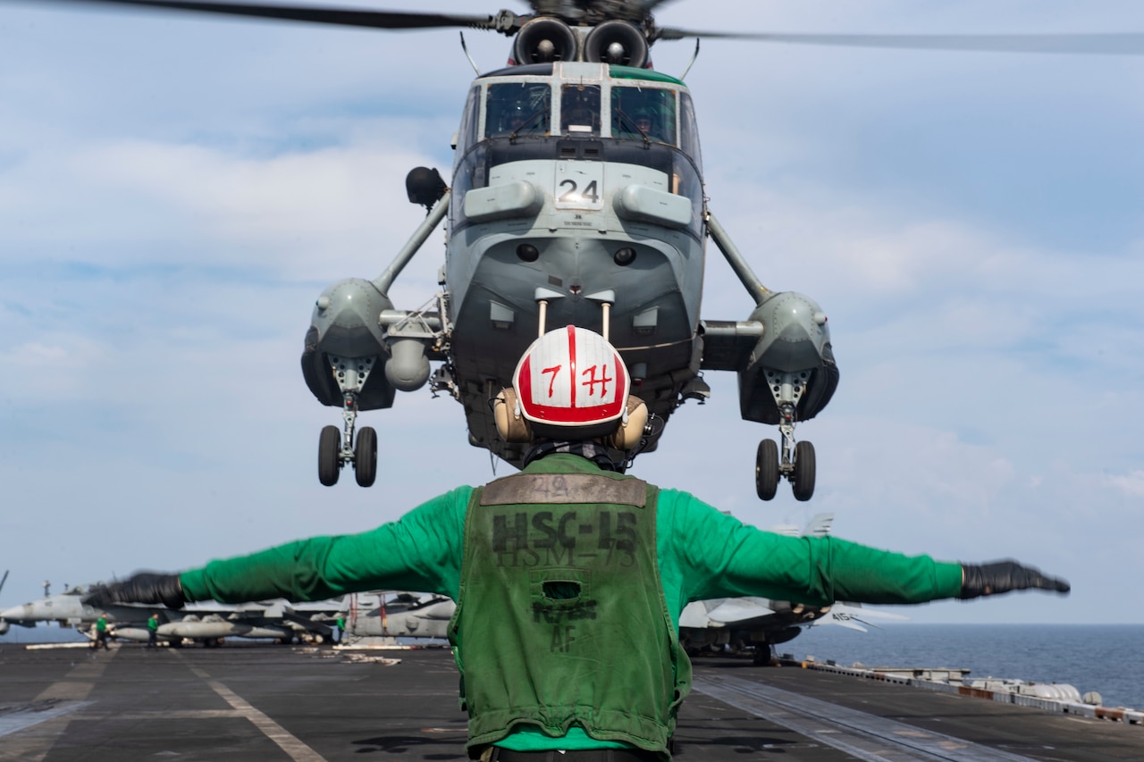 A sailor guides a helicopter as it lands on an aircraft carrier.