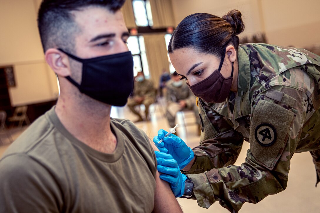 A female soldier wearing a face mask and gloves leans over to give an injection to a male soldier, who is seated.