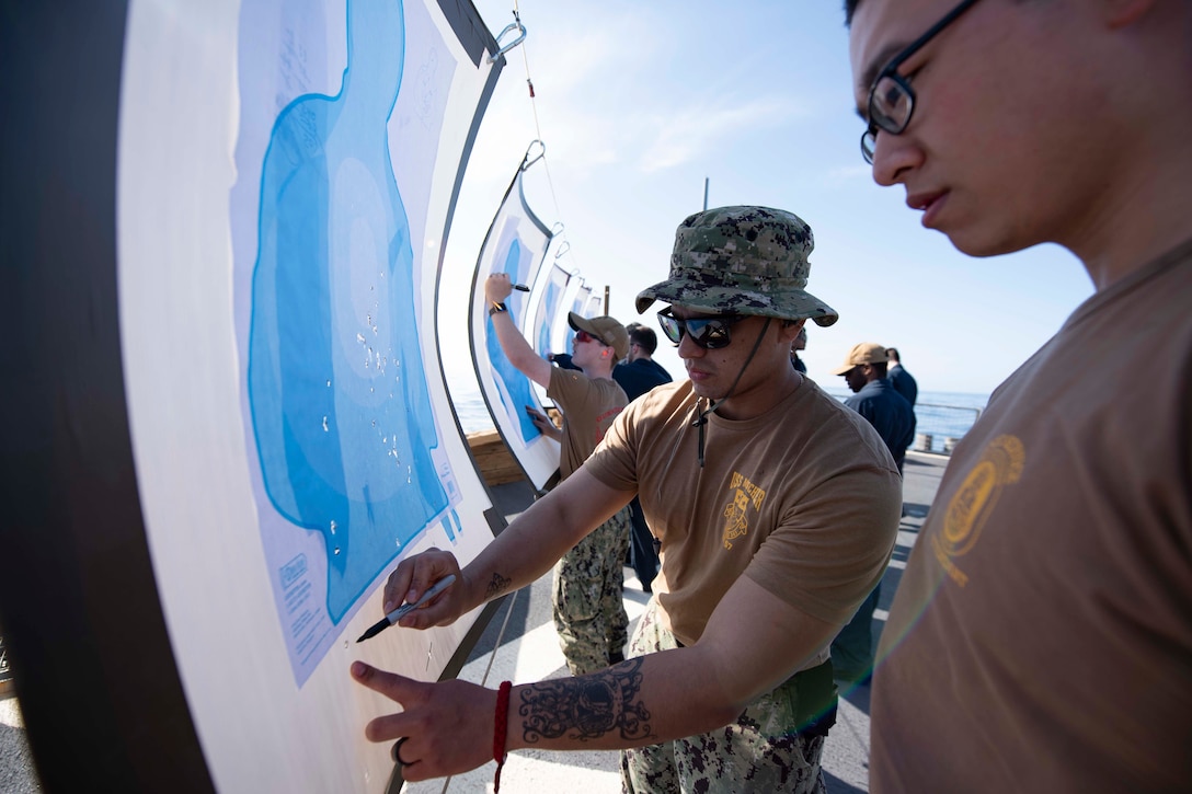 A group of sailors stand in a line and write on large sheets of paper with targets on them.