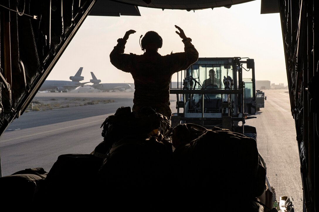 An airmen standing in an aircraft directs other service members as shown in silhouette.