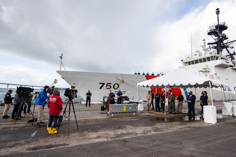 A man stands at a lectern set up outside and speaks to news crews gathered on a pier; a large ship is docked in the background.