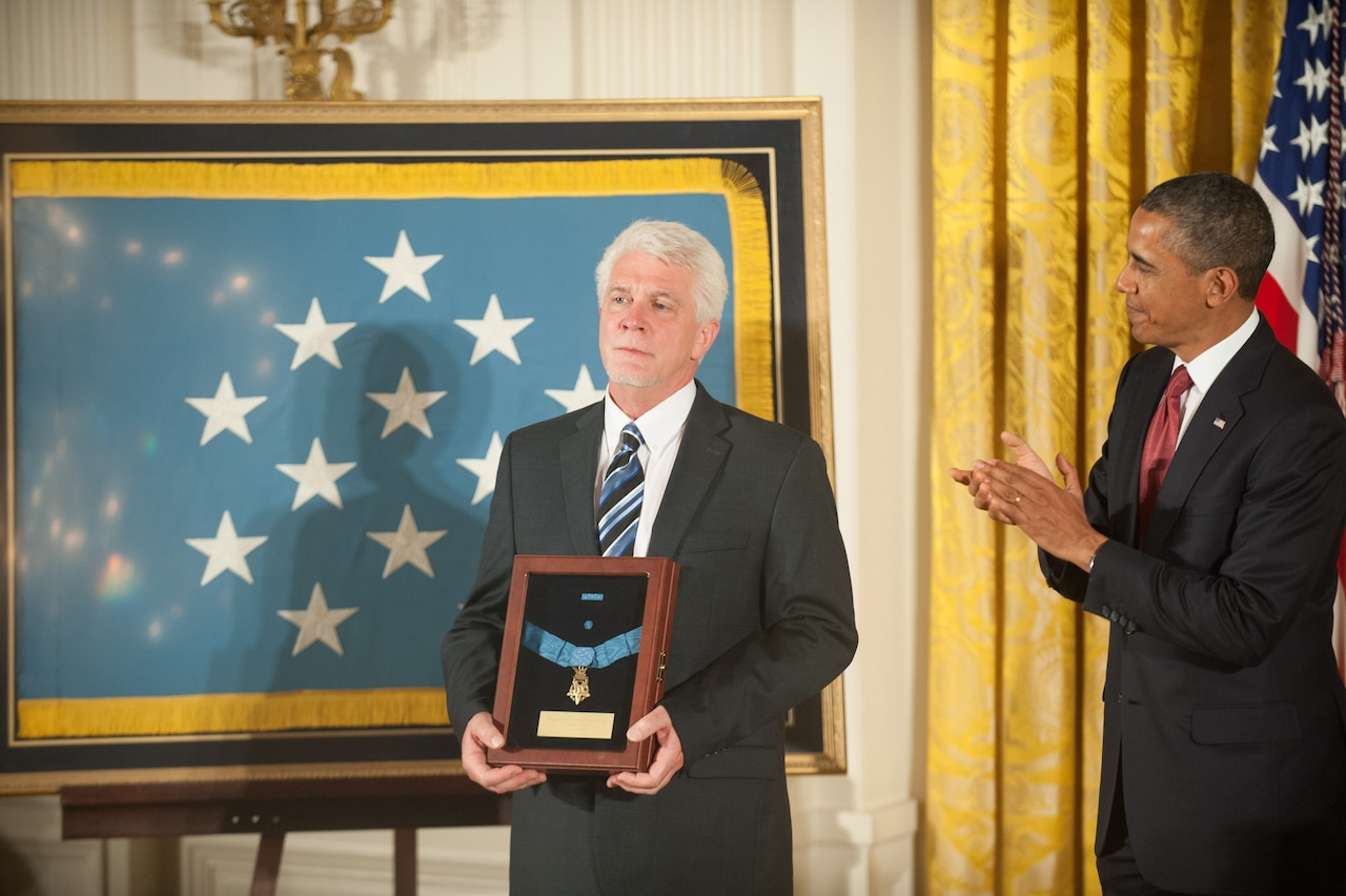 A man holds a shadow box with a medal as another man applauds beside him.