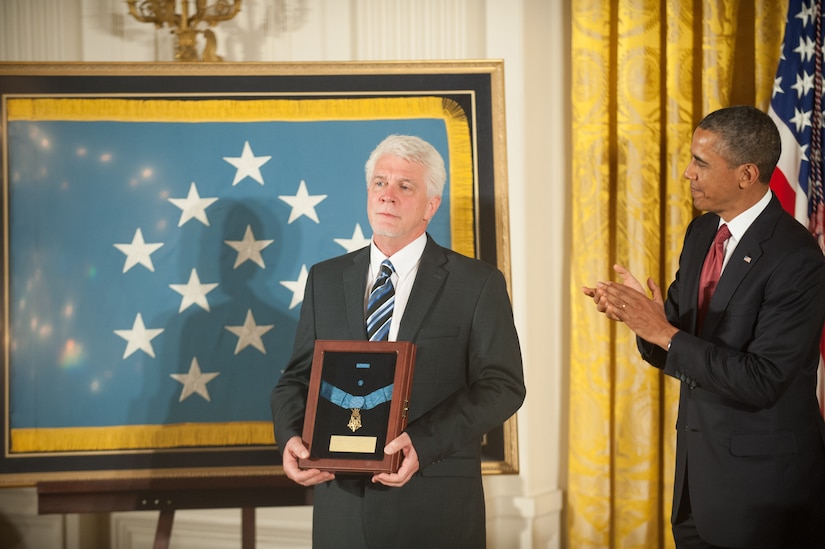 A man holds a shadow box with a medal as another man applauds beside him.