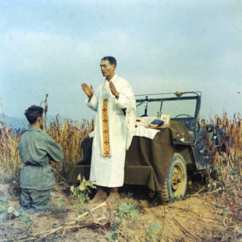 A priest blesses a kneeling man beside a Jeep in a field.