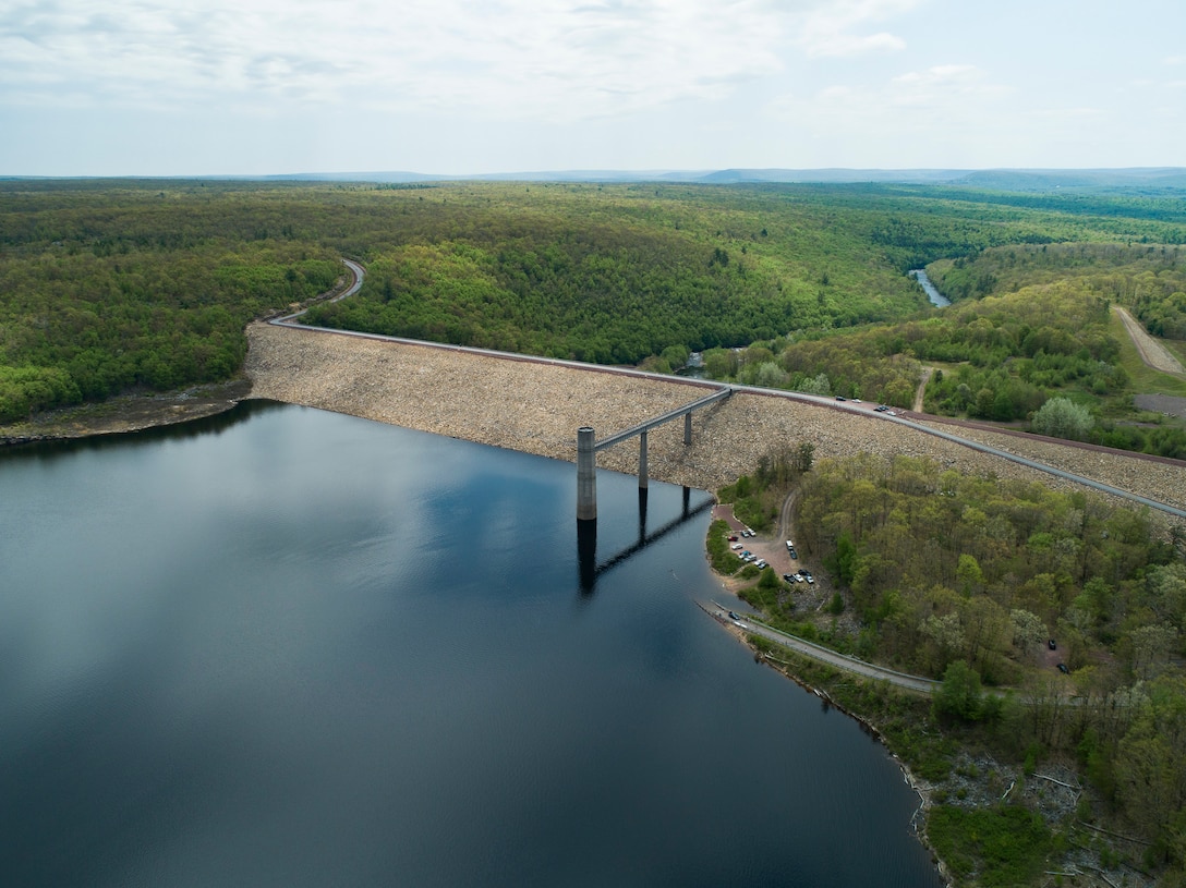 A large reservoir of water is pictured in front of a concrete control tower and dam with scenic vista of a creek/river valley in back drop of image.
