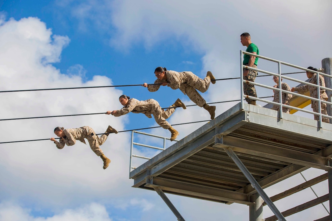 Three Marines Corps recruits slide along a rope suspended high above the ground as others watch and wait.