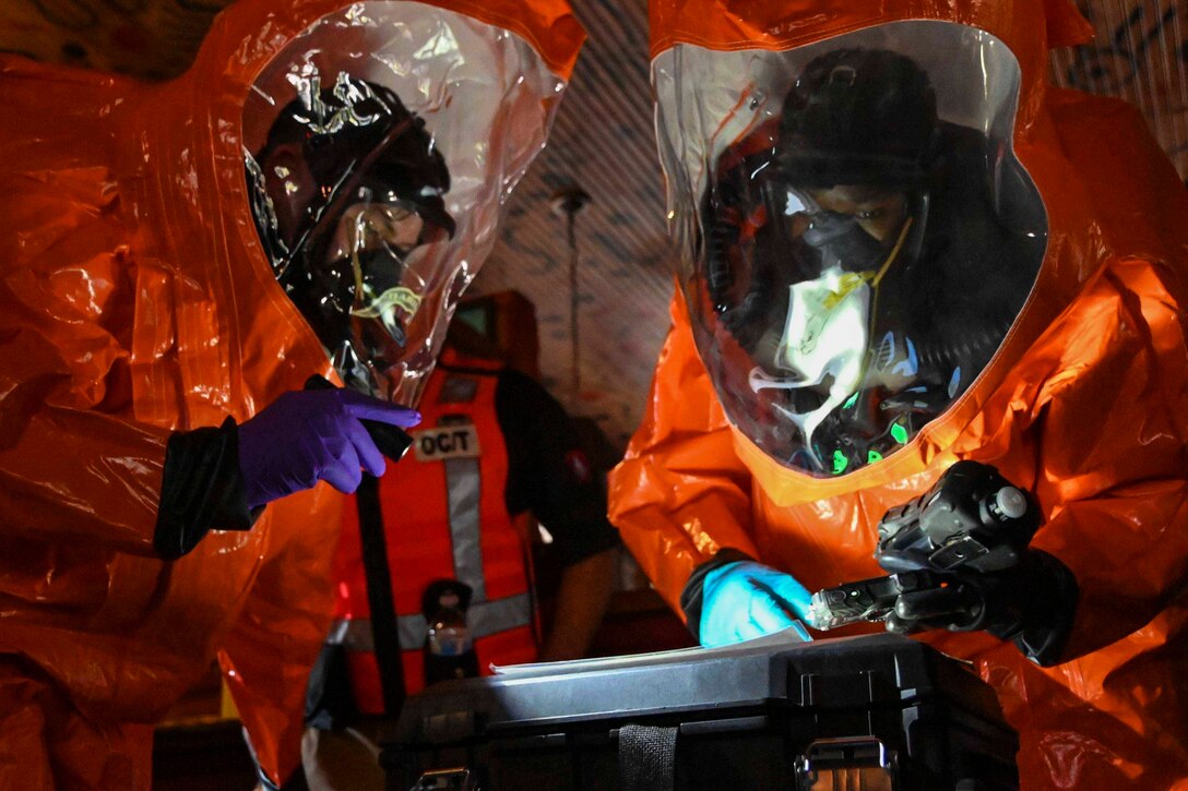 Guardsmen in protective gear use a flashlight to look at equipment.