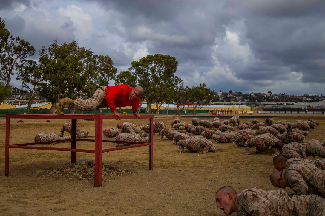 A large group of Marine Corps recruits do pushups on the ground while one Marine uses a raised platform.
