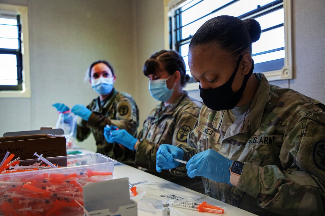Three soldiers wearing face masks and gloves prepare doses of the COVID-19 vaccine.