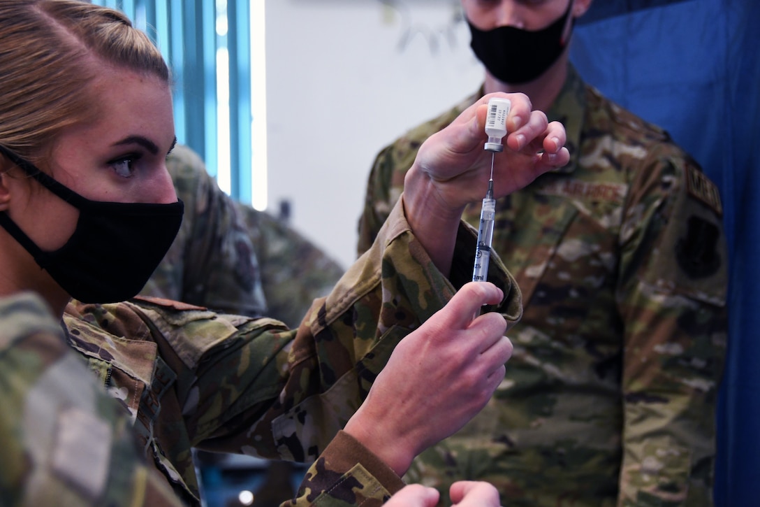 A soldier wearing a face mask holds a syringe while inserting the needle into a vial.