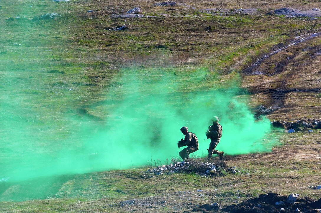 Soldiers fire weapons in a field surrounded by a cloud of bluish green smoke.