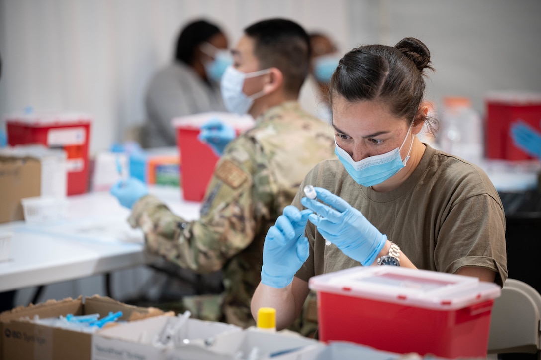 A soldier wearing a face mask and gloves holds a syringe while inserting the needle into a vial to prepare for a  COVID-19 vaccination.