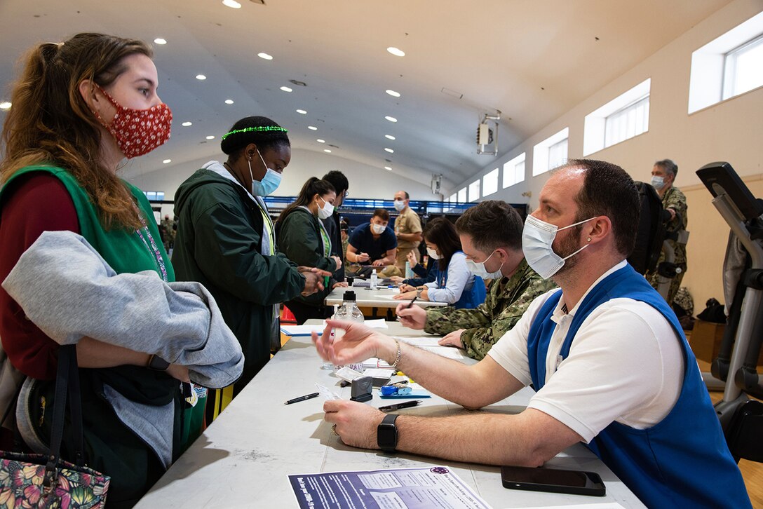 Civilian and military personnel wearing face masks sit at a table to participate in a COVID-19 vaccine distribution exercise for essential front line personnel.