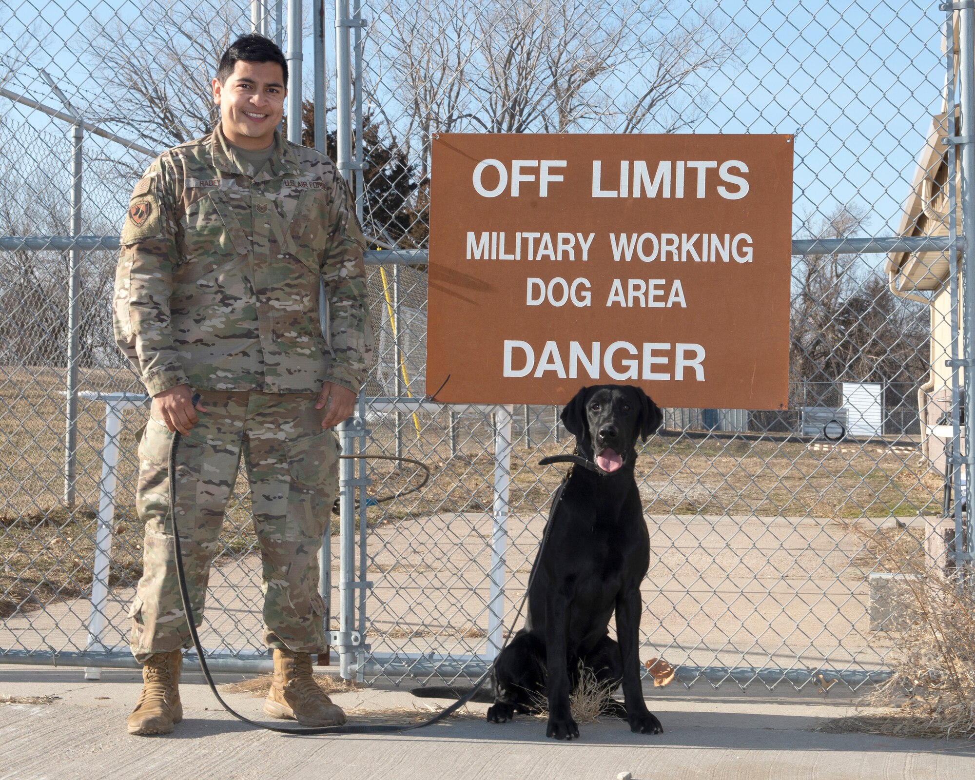 Air Force sergeant standing in front of brown sign on fence that reads off limits military working dog area danger, military working dog, black labrador retriever sits squarely underneath the sign