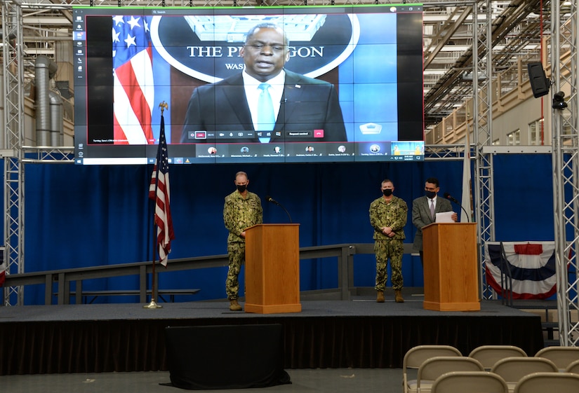 Two military personnel and a man in civilian clothes stand behind lecterns on a stage. In the rear, a large display screen shows a video that features the secretary of defense speaking from the Pentagon..