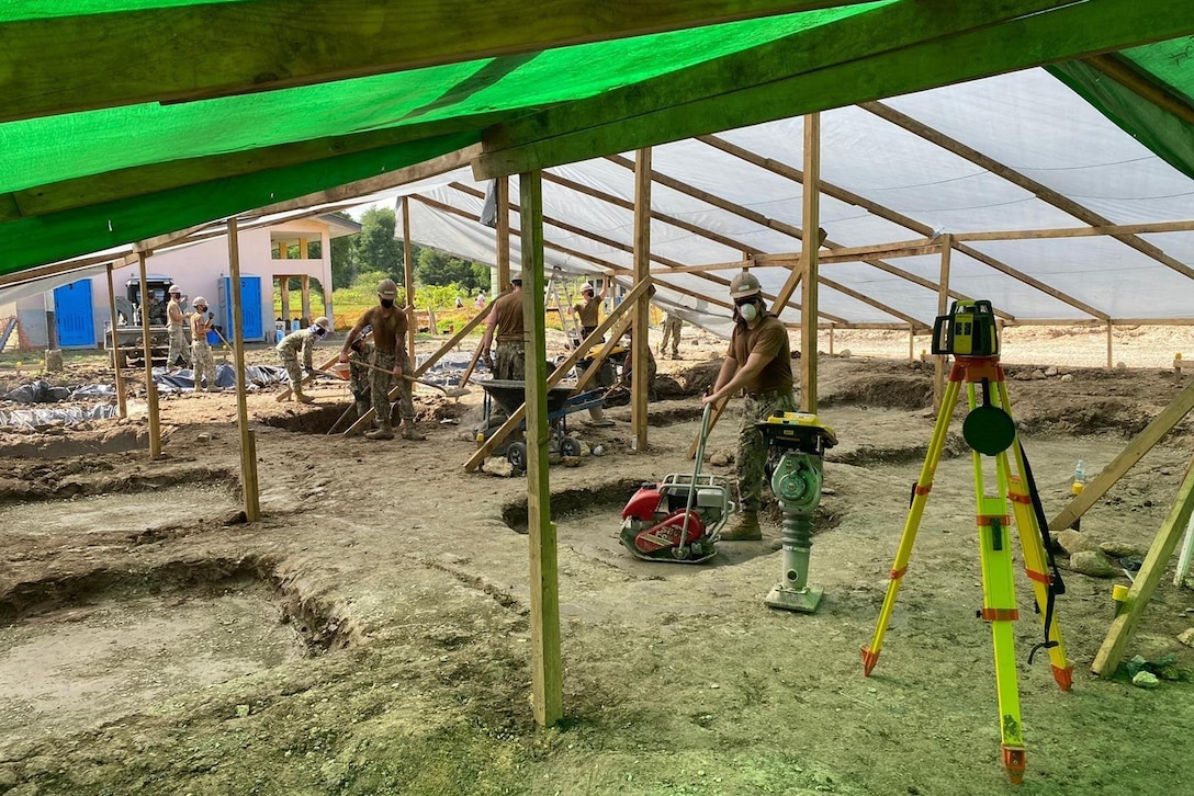 A group of men use construction equipment under a large green tarp.