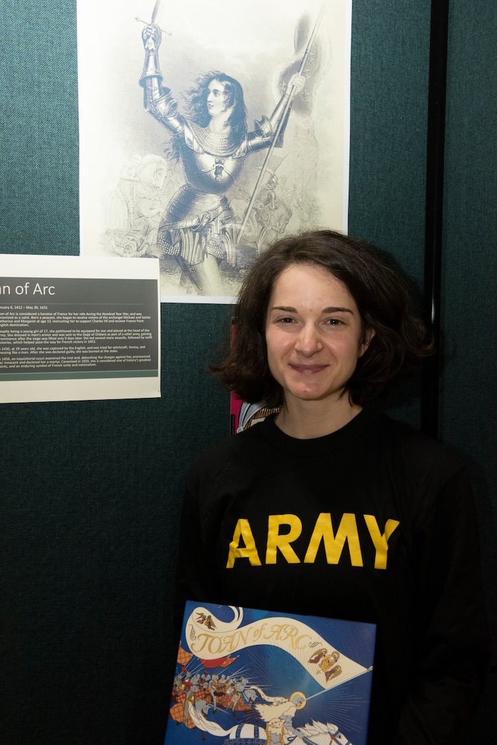 Army Staff Sgt. Samantha Fontaine poses for a photo in front of a display of Joan of Arc in the Green Mountain Armory at Camp Johnson, Vermont, March 18, 2021. Fontaine, a retirement services NCO with Joint Force Headquarters - Vermont National Guard, was one of several Guardswomen who facilitated a book reading and display of Guard uniforms and equipment via teleconference for a local day care center. (U.S. Army National Guard photo by Don Branum)