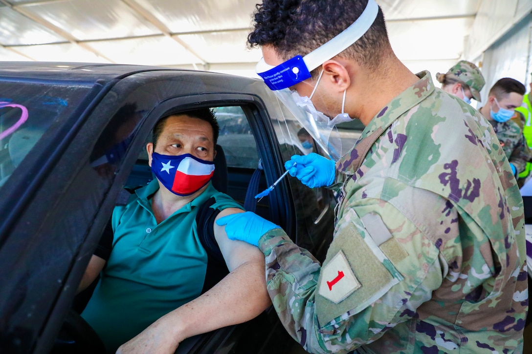 A soldier wearing a face mask and gloves gives a COVID-19 vaccine to a man seated in a vehicle wearing a face mask.
