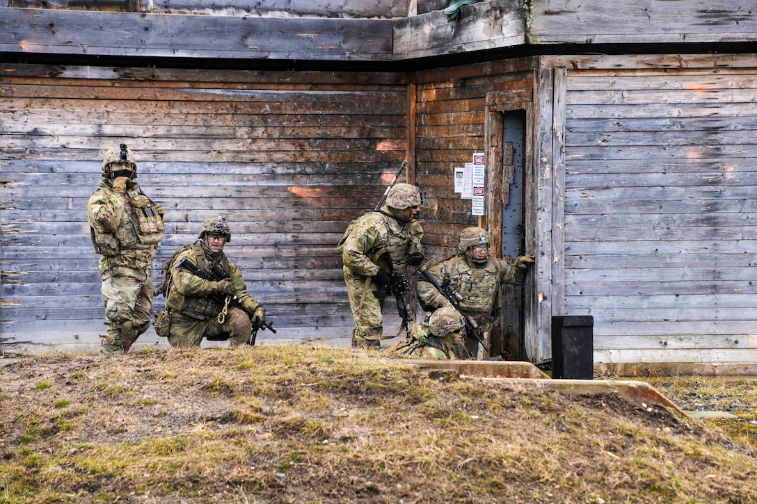Five soldiers stand and kneel behind the wall of a building.