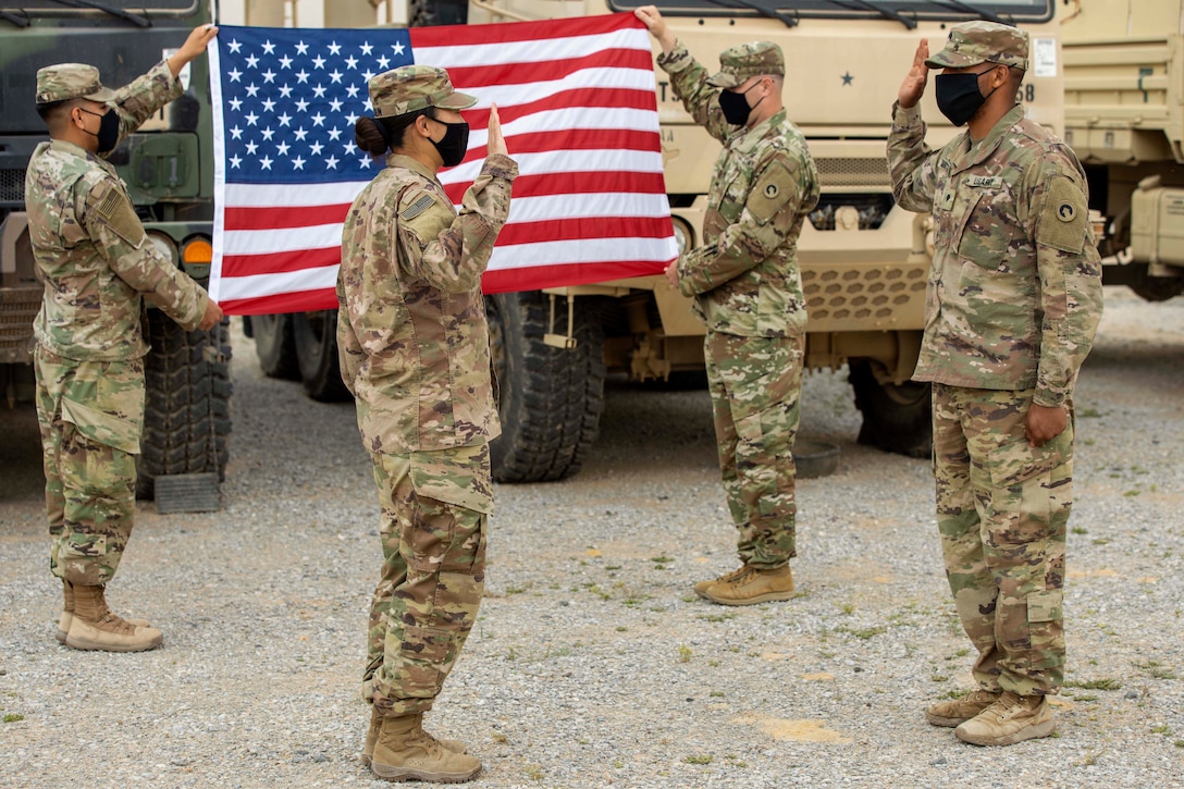 Two soldiers hold up their right hands while facing each other in front of an American flag.
