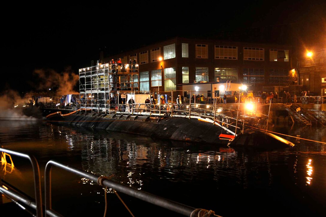 At night, a submarine sits submerged in the water in front of an industrial complex.