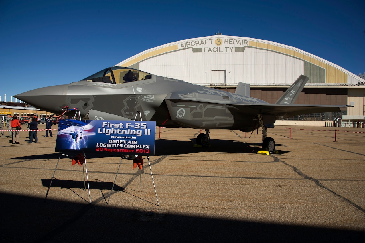 A military fighter jet sits outside a hangar. A paper sign in front of the aircraft reads "First F-35 Lightning II."