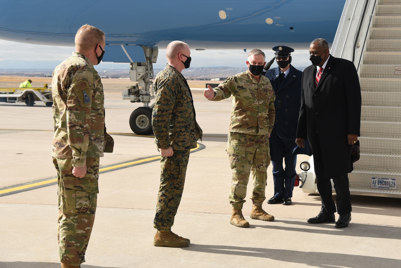 Three service members stand in a row to greet the Secretary of Defense, who has disembarked from a plane.