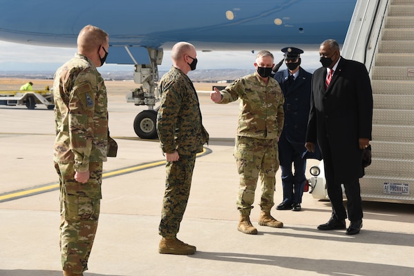 Three service members stand in a row to greet the Secretary of Defense, who has disembarked from a plane.