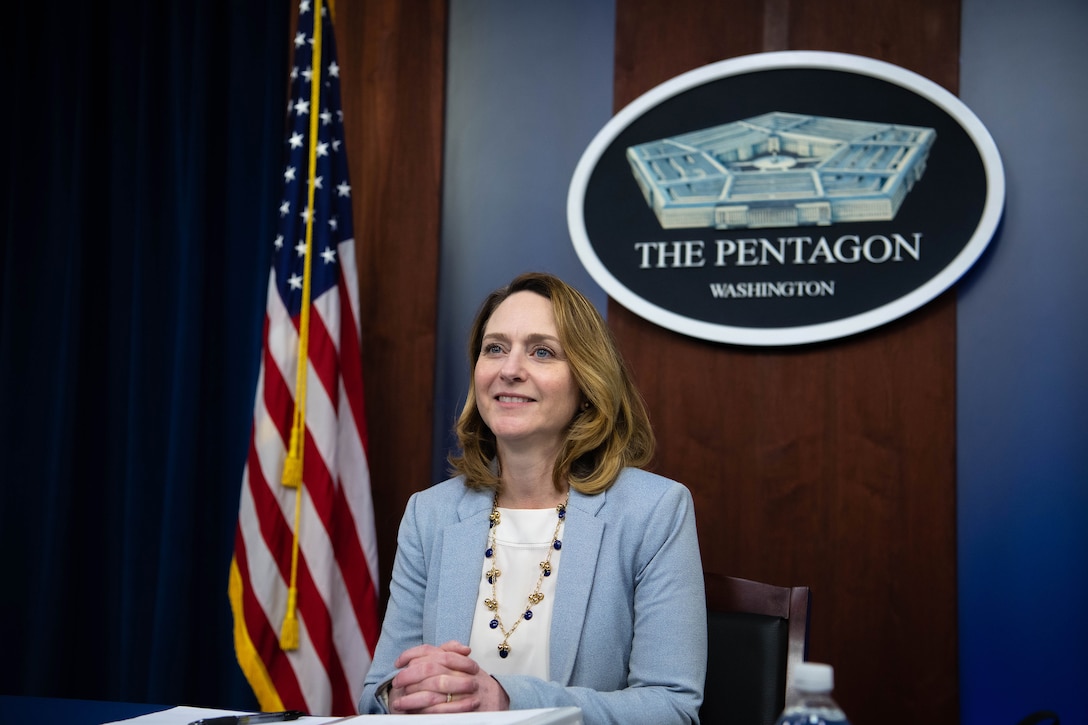 Deputy Defense Secretary Kathleen H. Hicks sits at a table, with a Pentagon seal displayed on the wall behind her.