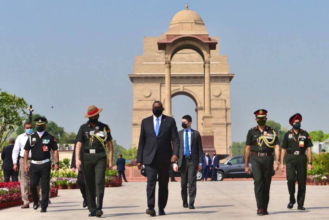 A group of men, some in military uniforms, walk toward the camera. The India Gate is behind them.