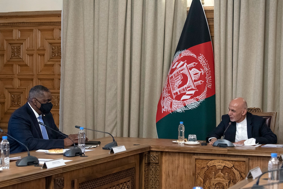 Secretary of Defense Lloyd J. Austin III talks with another civilian at a table in front of an Afghan flag.