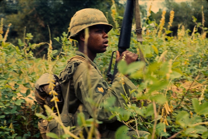 A soldier holding a weapon up kneels in vegetation.