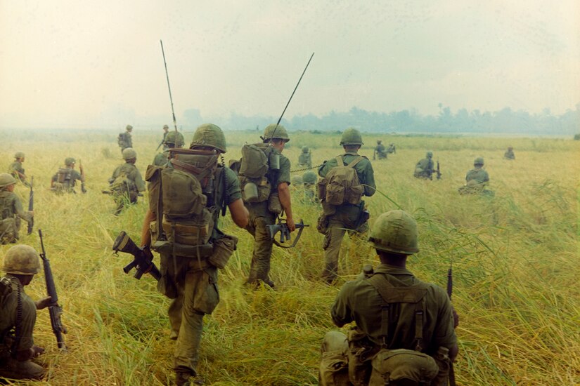 Soldiers wearing olive-colored uniforms and carrying weapons, shown from behind, kneel or walk across a rice field.